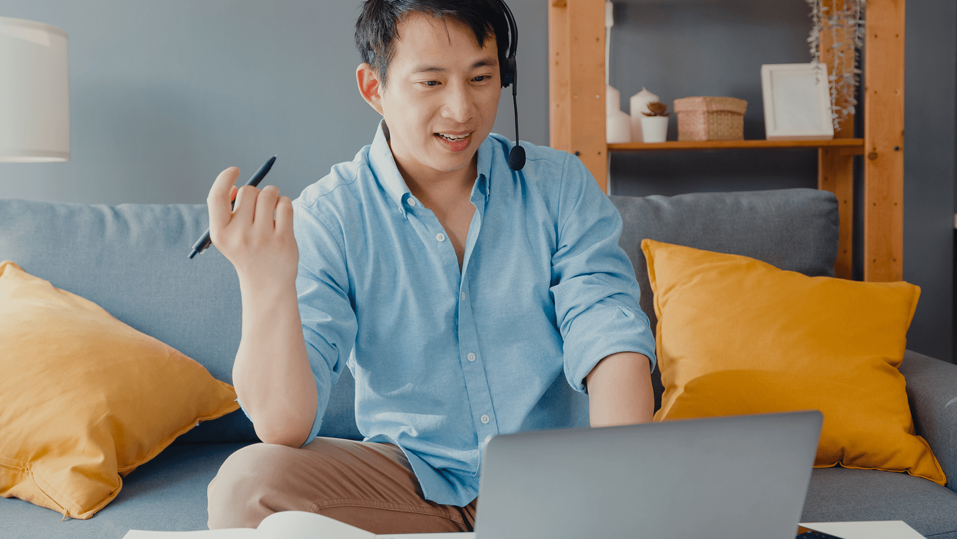 A focused man works from home, engaged in a conversation through his headset while gesturing with his pen, with his laptop open in front of him and notepads on the coffee table, all set against a cozy living room backdrop with a blue couch and bright yellow cushions.