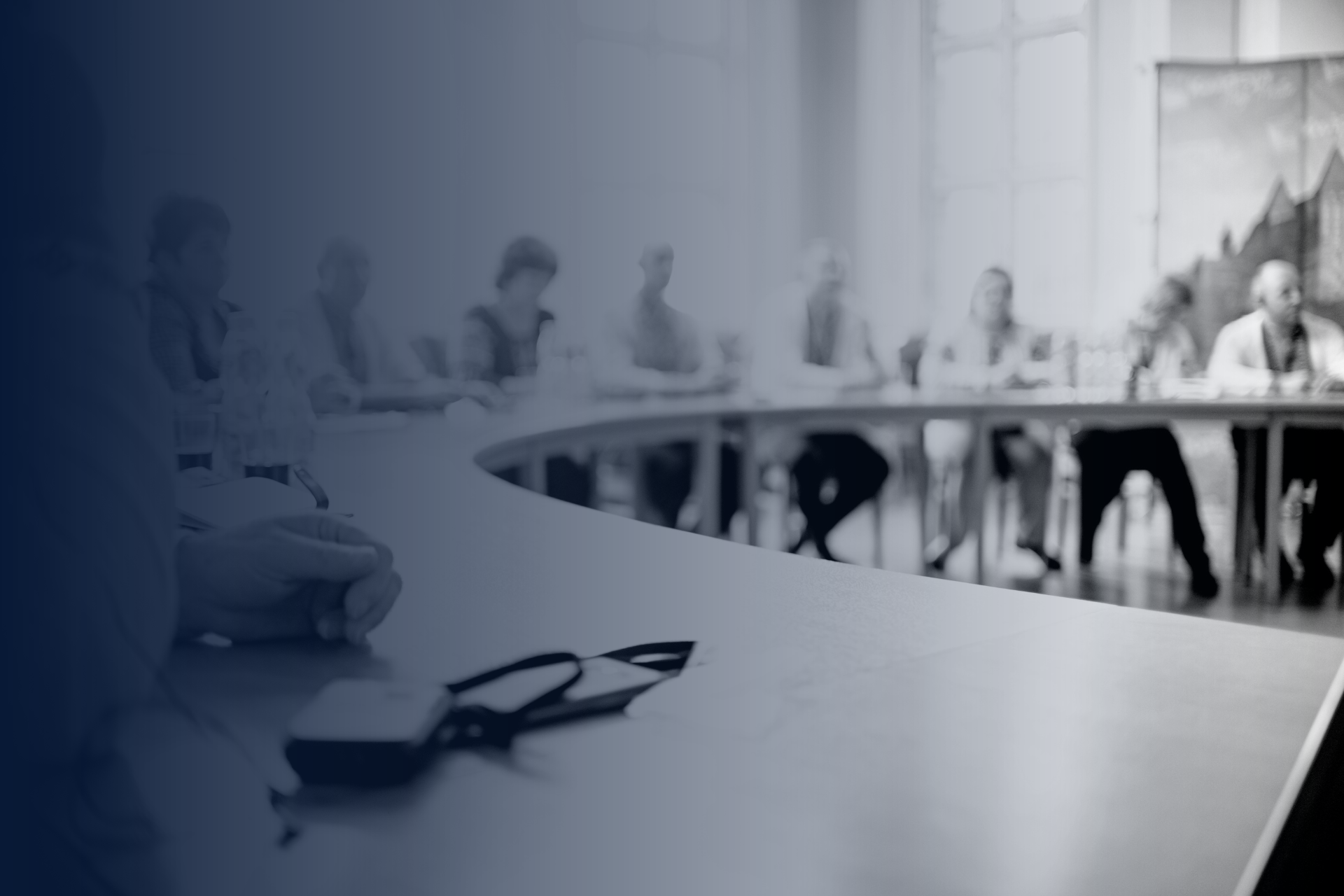 Monochrome image of a blurry boardroom meeting with attendees seated around a long table, suggesting a focus on corporate engagement or discussion.