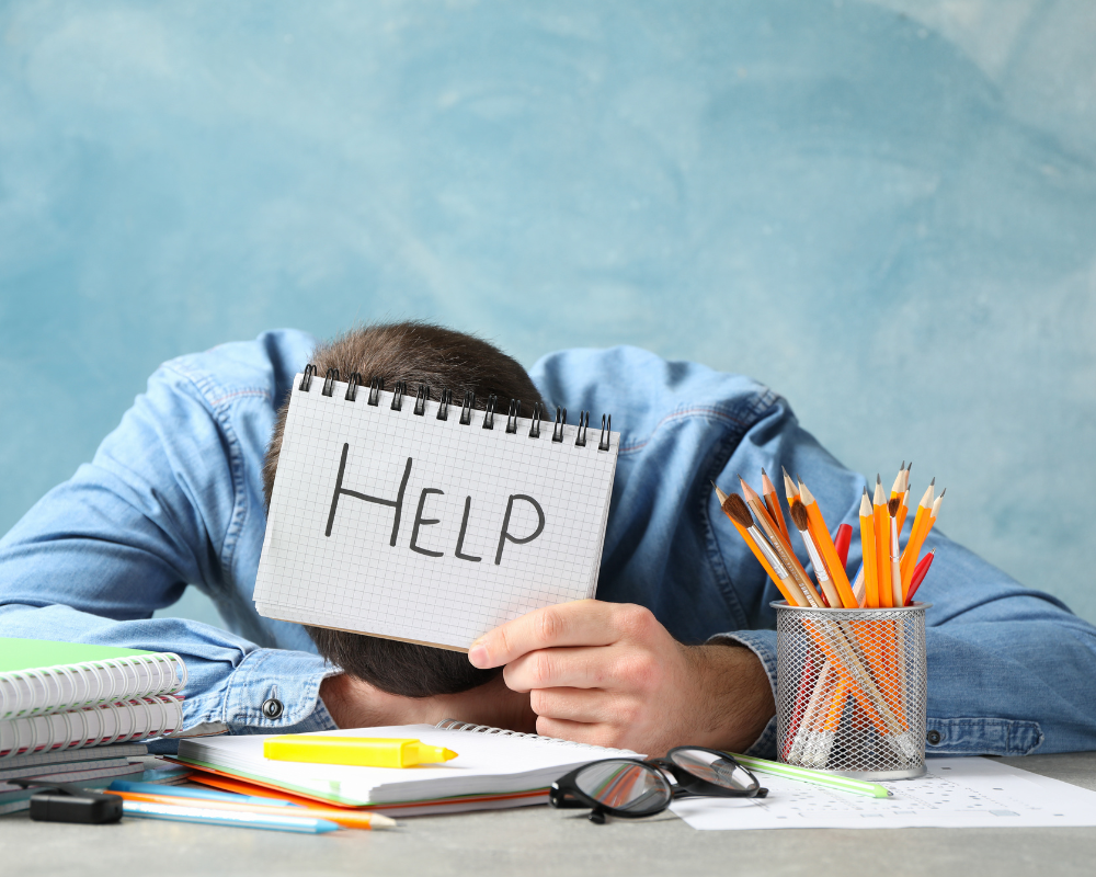 A man with his head on the desk holding a sign asking for help