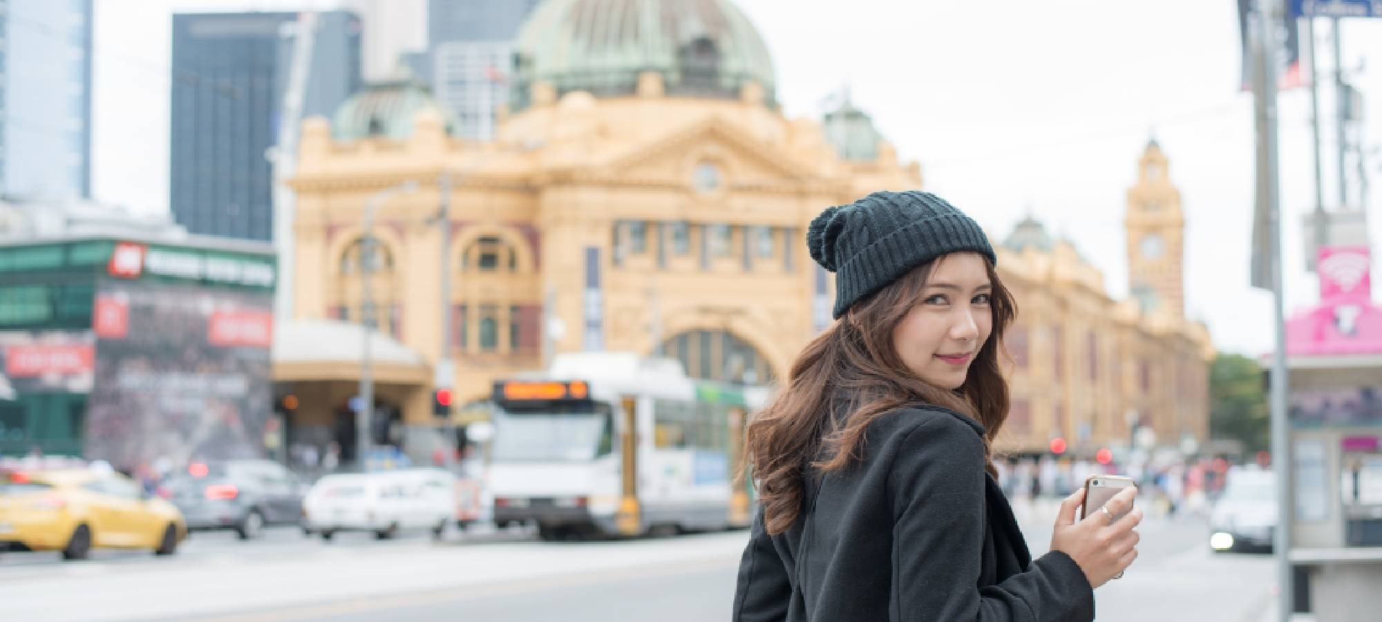 Person in a beanie holding a phone and looking back outside Flinders street station in Melbourne. 