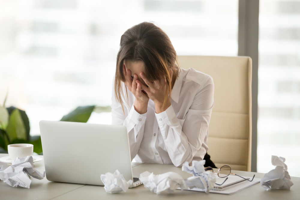 Stressed tired businesswoman feels exhausted sitting at office desk with laptop and crumpled paper.