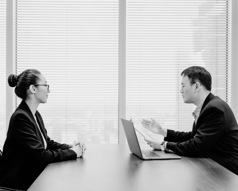 Black and white image of a job interview in progress, with a candidate and recruiter seated across from each other at a desk, discussing opportunities.