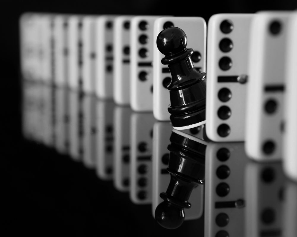 A black pawn standing firm against a row of falling dominoes, symbolizing stability and strategic positioning amid disruption.