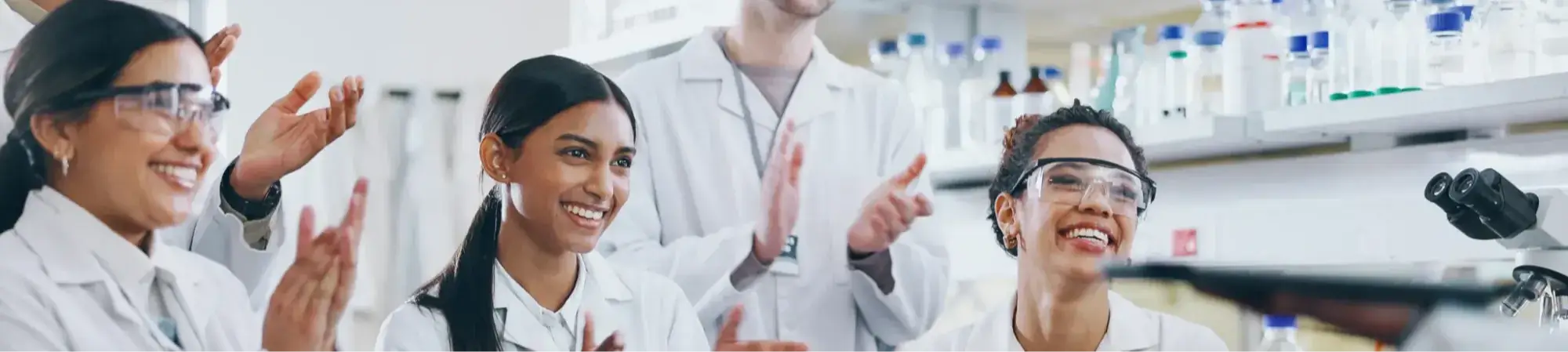 A diverse group of scientists in white lab coats and safety goggles smiling and clapping in a modern laboratory.