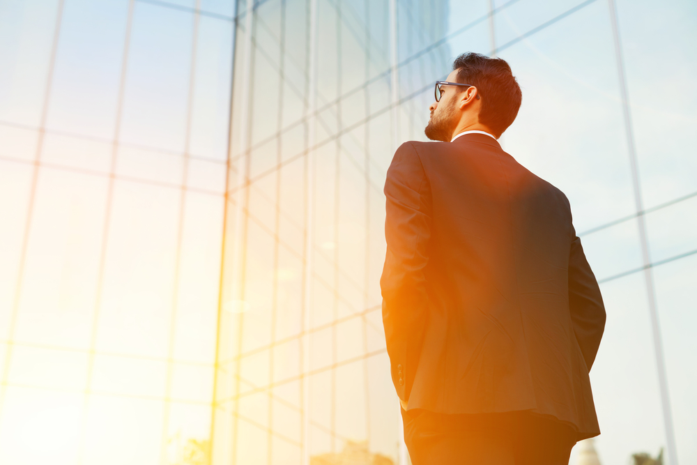 Back view of rich businessman with hands in pockets looking on his skyscraper building while standing outside