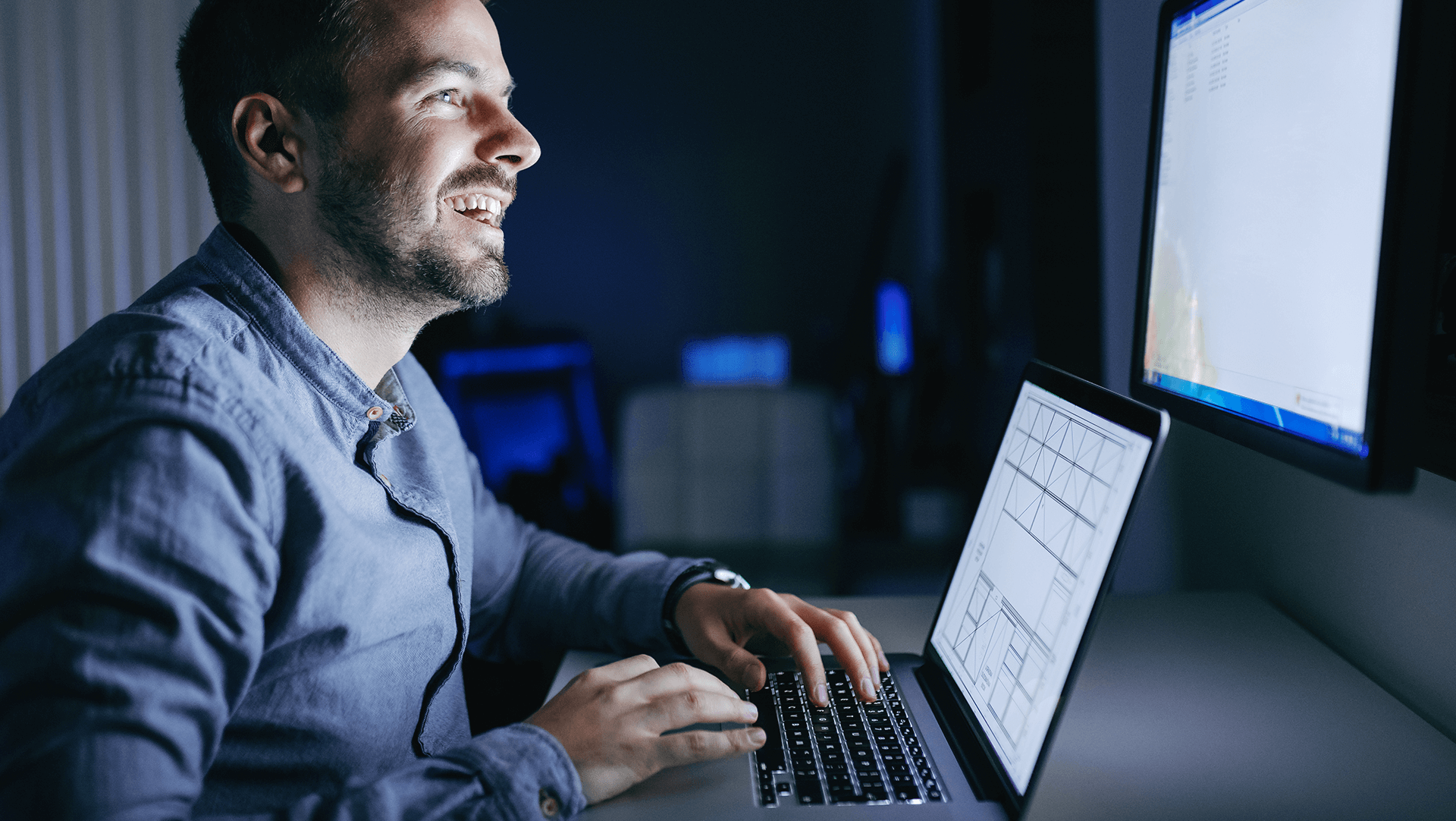 Man smiling as he works on a laptop in a dimly lit room with a computer monitor in the background.