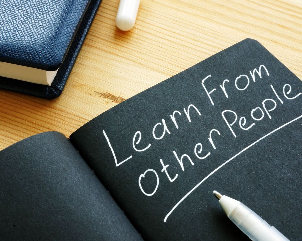 The phrase ‘Learn from other people’ written with a white pen on black paper, placed on a desk, symbolising knowledge-sharing and learning from others’ experiences.