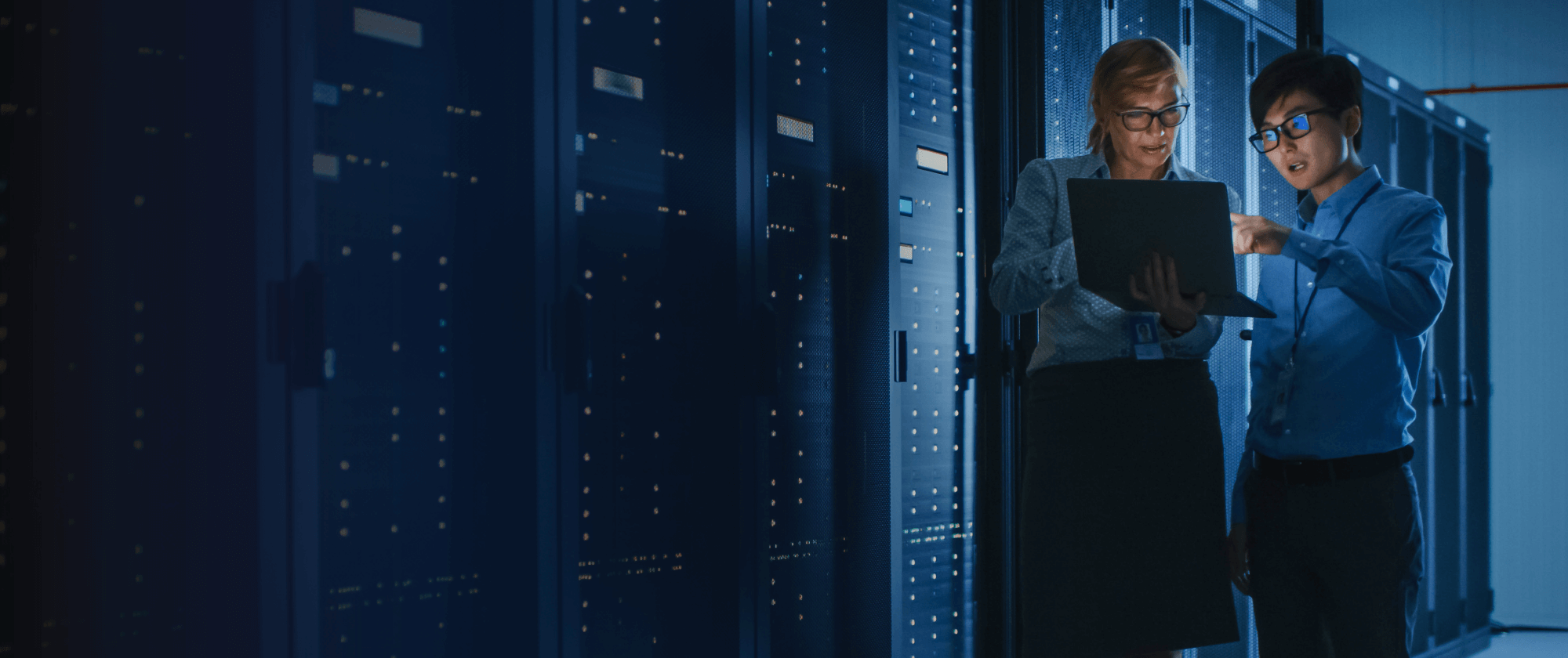 Two it professionals analyzing data on a laptop in a server room at night.