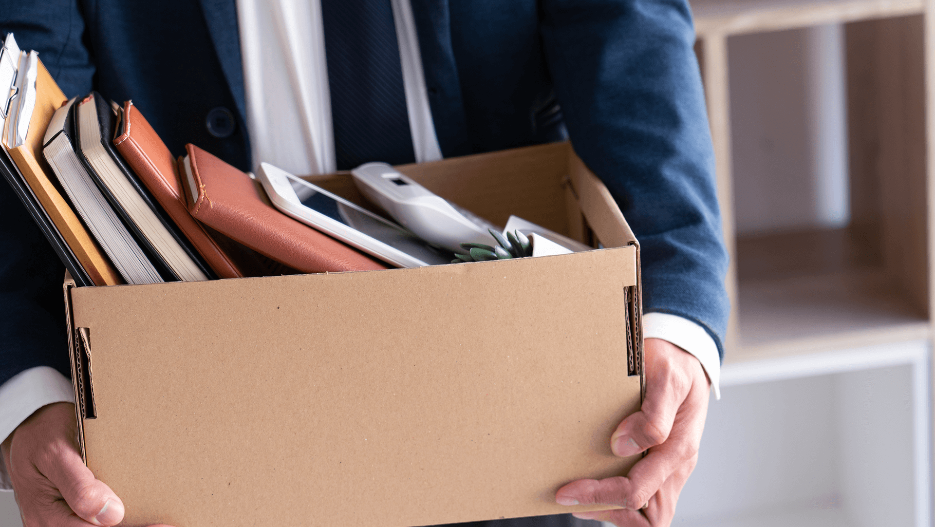 A person in a suit carrying a cardboard box full of work-related items, possibly indicating a job change or office relocation.