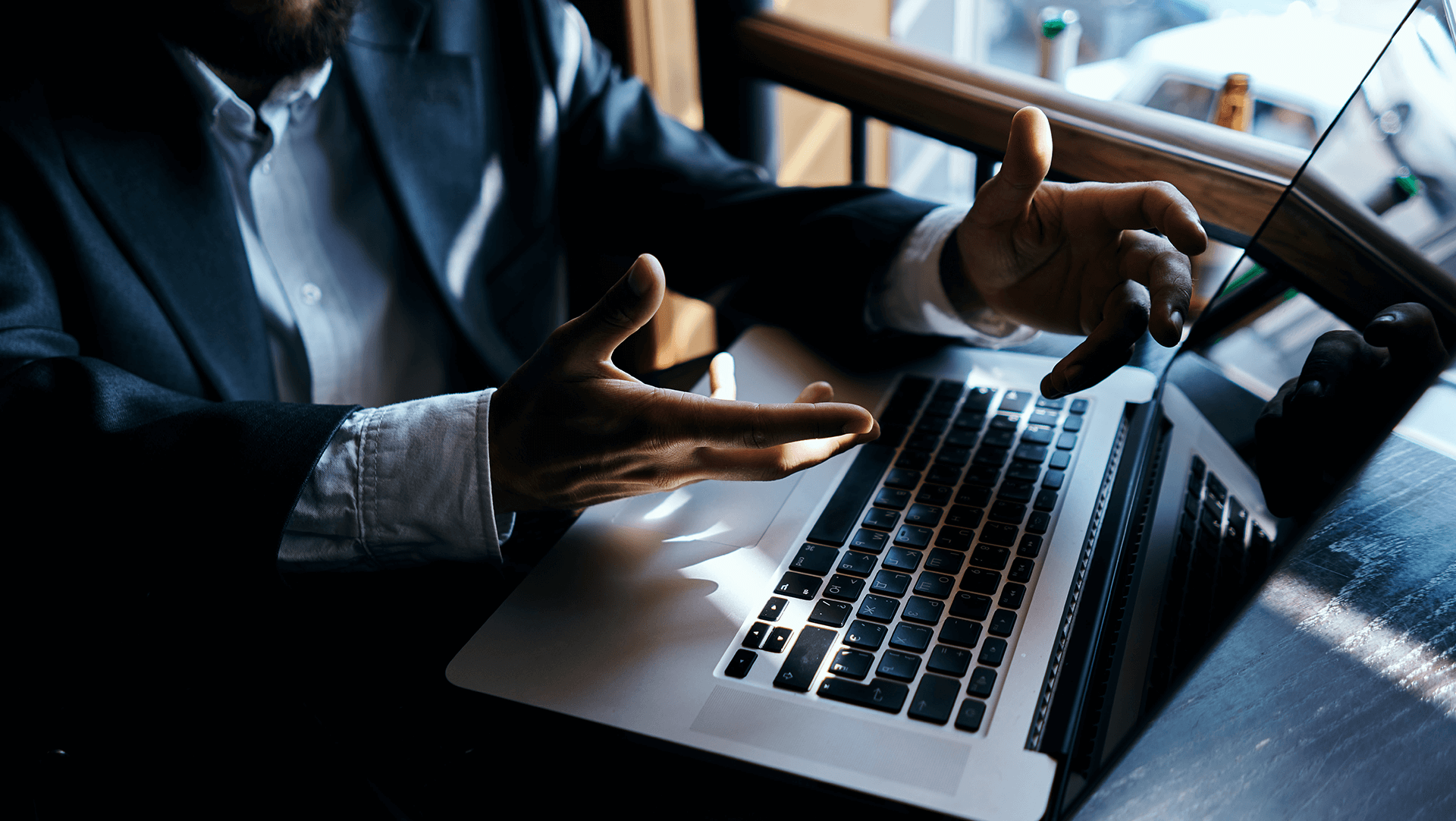 A person in a business suit gesturing with hands during a video call on a laptop in a dimly lit office setting.