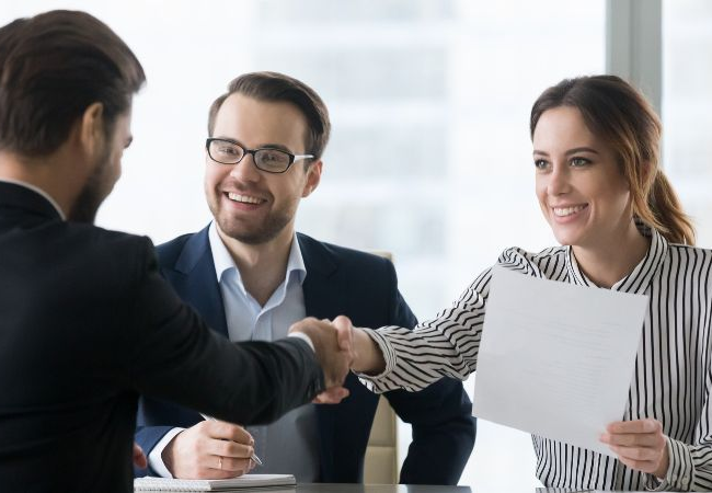 Woman shaking hands with a man across the desk with another man looking on. 