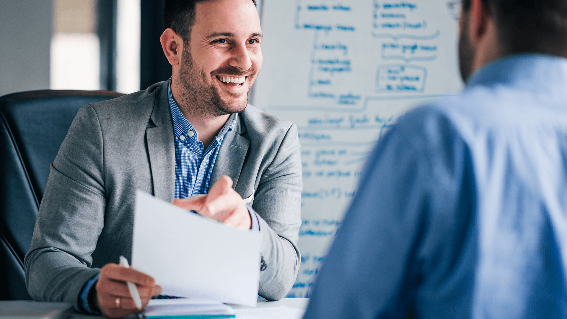 Two professionals in a meeting, with one handing over a document to the other, sharing a moment of lighthearted interaction in a bright office setting.