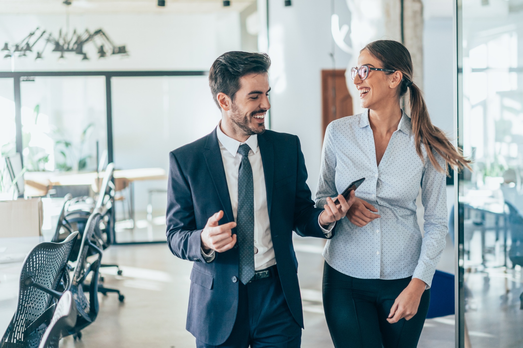Two coworkers smiling while looking at a phone outside an office room.